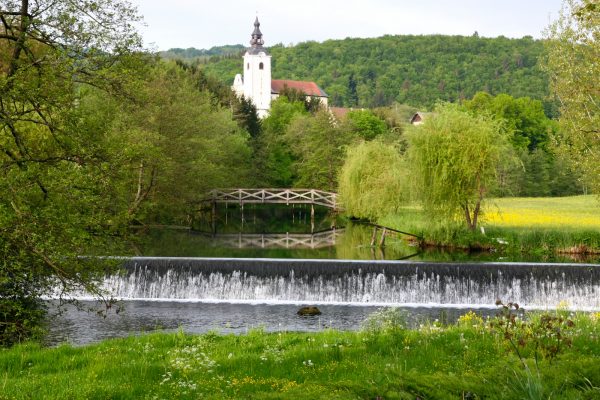 Pogled na cerkev, lesen most čez reko in jez v zeleni gozdnati pokrajini