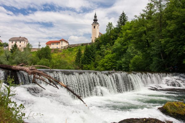 Cerkev z zvonikom nad širokim slapom in brzicami, obdana z zelenim gozdom